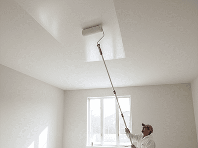 A paint roller on an extension pole applying white paint to a ceiling, working away from a window.