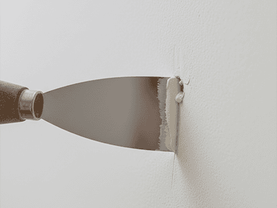 A close-up of a putty knife pressing white filler into a small hole on a gyprock wall.